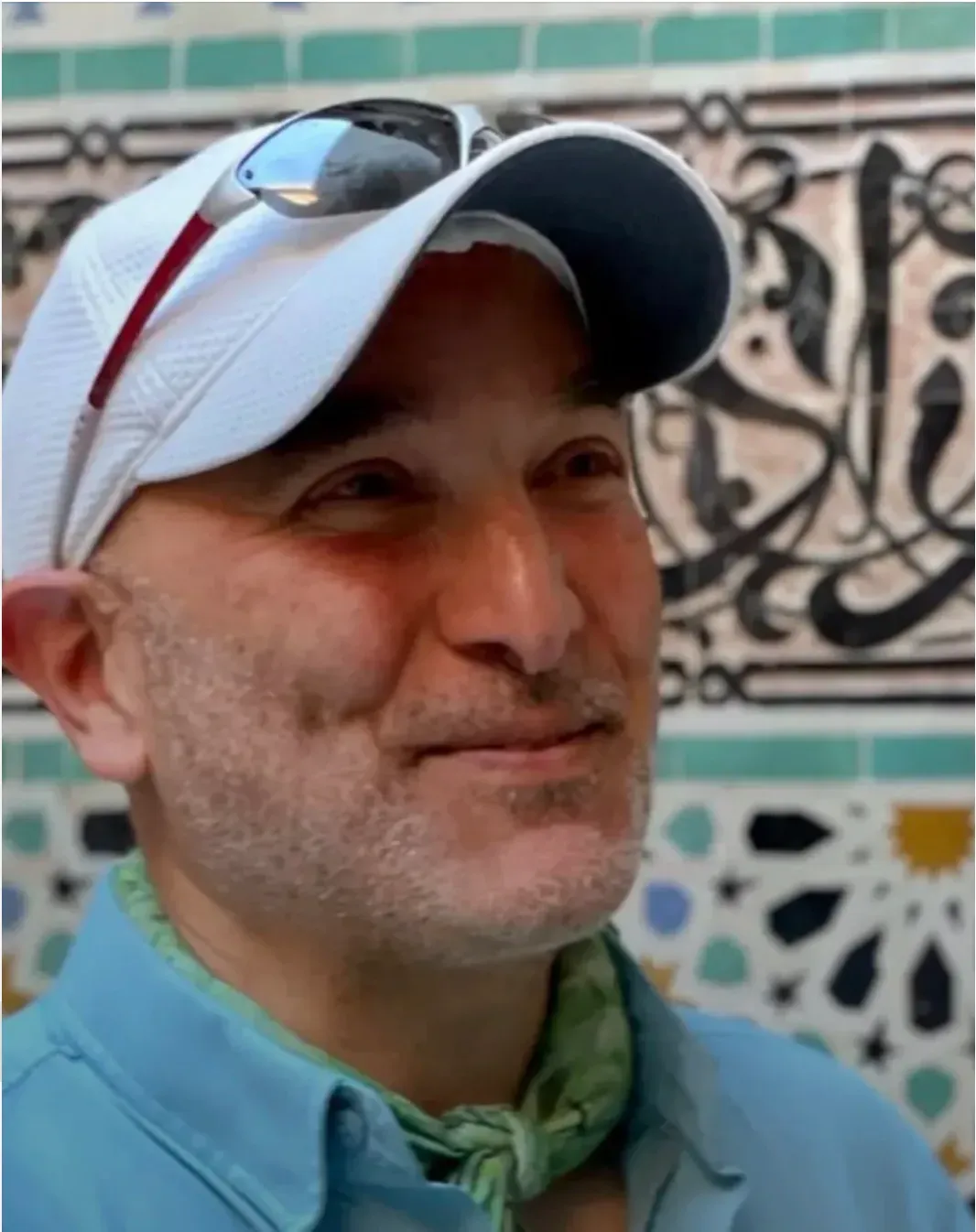 Man smiling in a white cap and blue shirt, standing before patterned tile wall