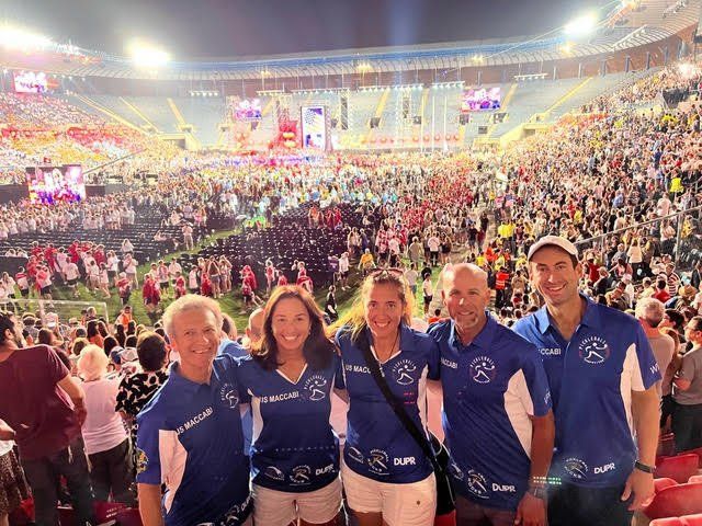 Five people in blue shirts posing at a brightly lit stadium with a large crowd in the background