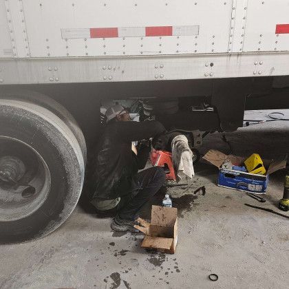 Mechanic under a white truck, working near the wheel in a garage with tools and parts scattered nearby