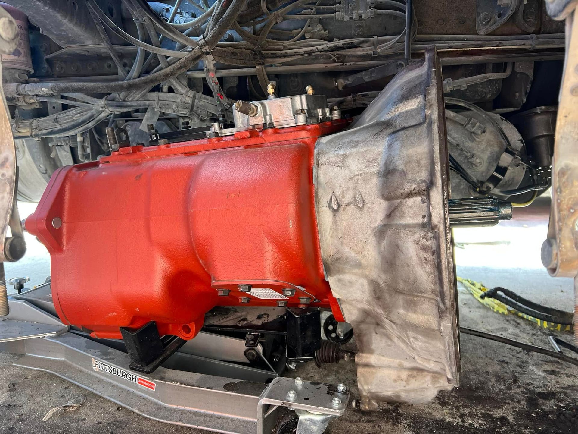 Red car transmission removed, hanging beneath the vehicle on a lift in a repair shop.