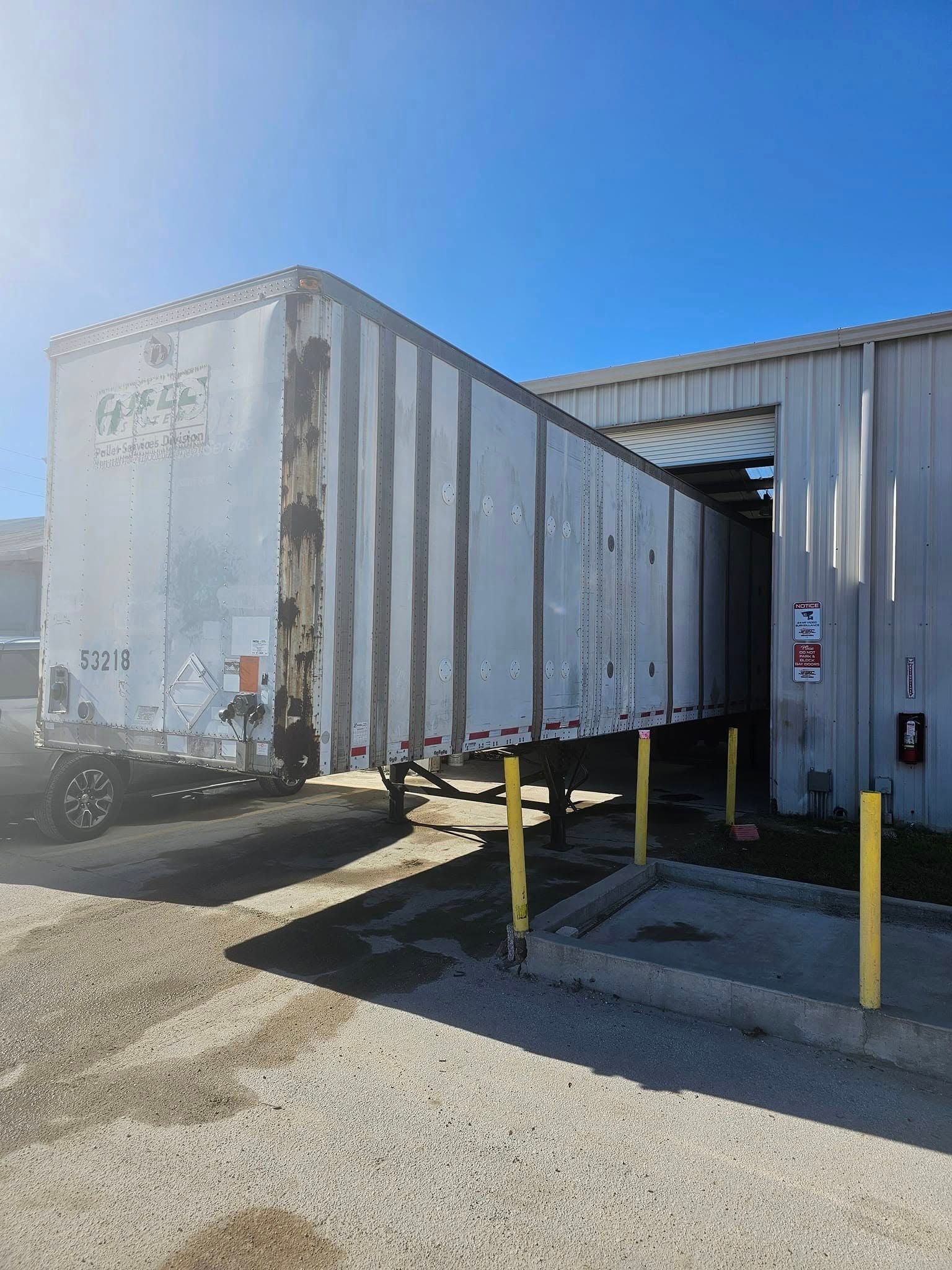 White trailer backed into a loading dock beside a warehouse, with yellow safety posts and a clear blue sky.
