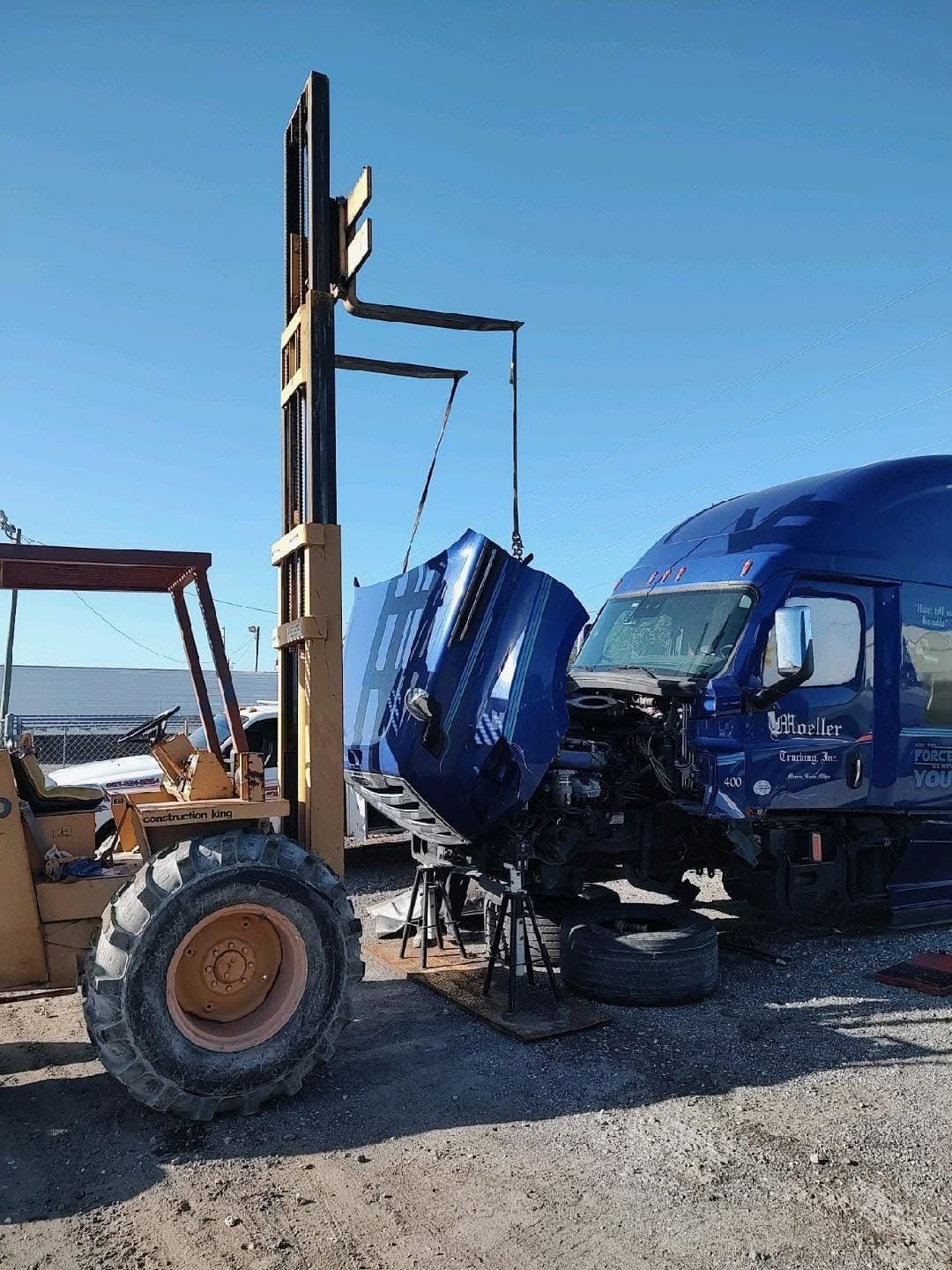 Forklift lifting a blue truck cab in an outdoor lot, with another blue truck nearby.