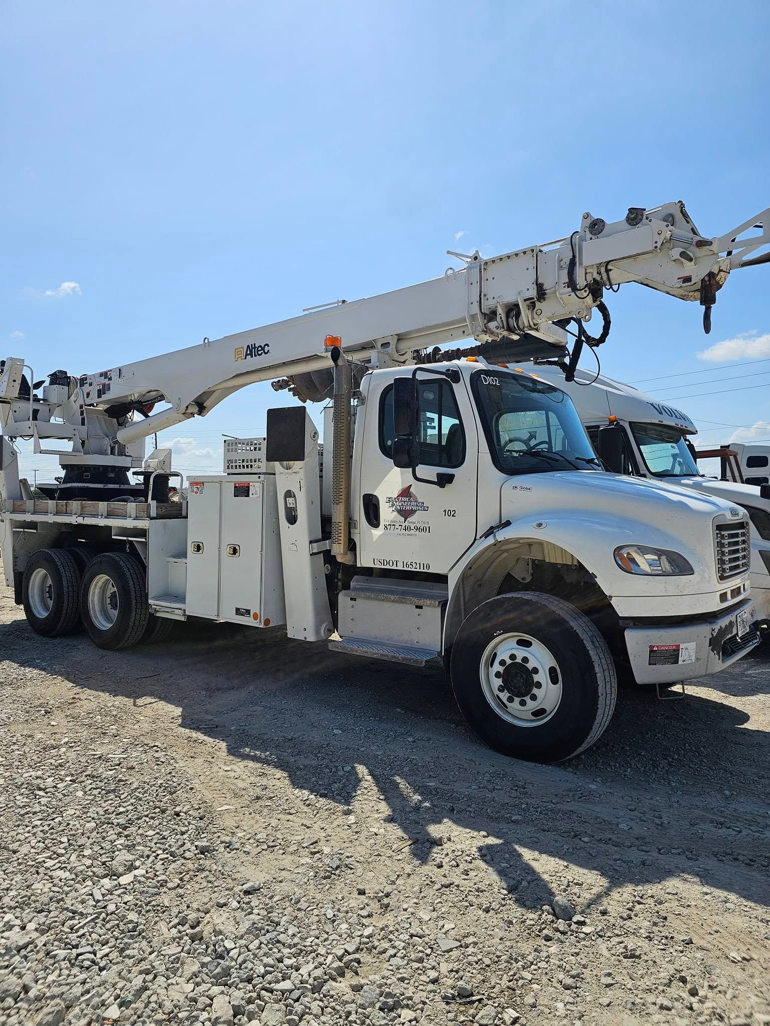 White utility truck with extended boom lift parked on gravel under a blue sky.