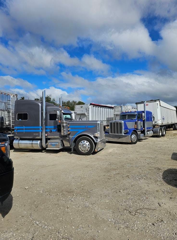 Black semi truck and trailers parked on a gravel lot under a cloudy blue sky.