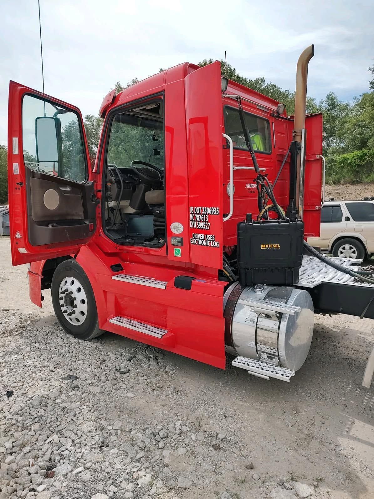 Red semi-truck cab with driver door open, parked on a gravel lot.