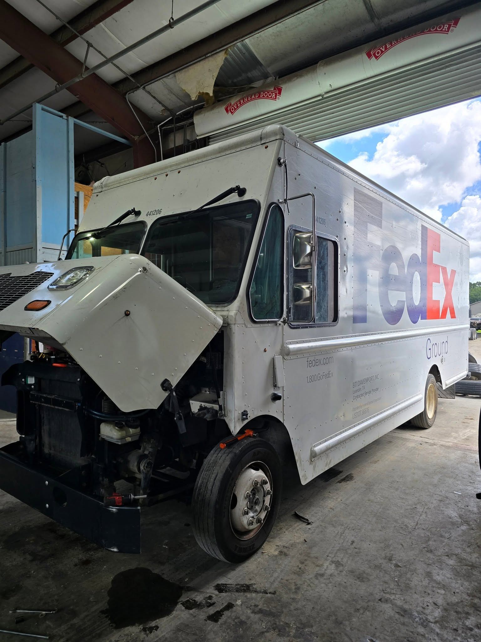 White delivery truck with open front hood in a garage bay, showing damage and repair work.