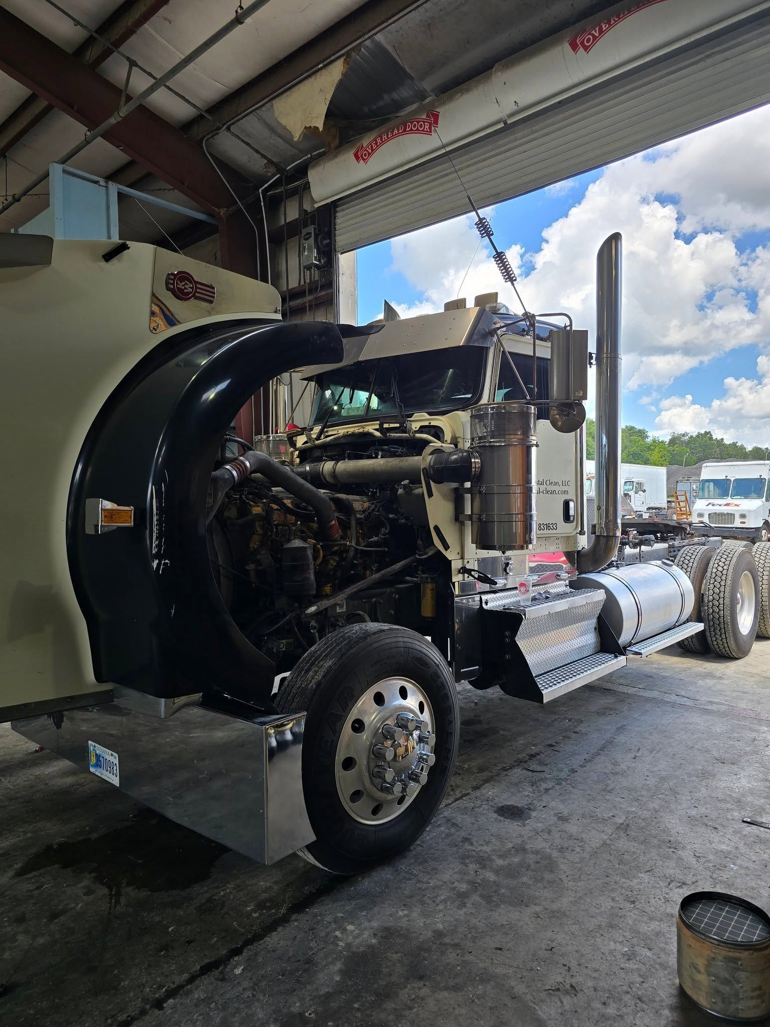Black semi-truck in a garage, hood open, showing engine and front wheels under repair.