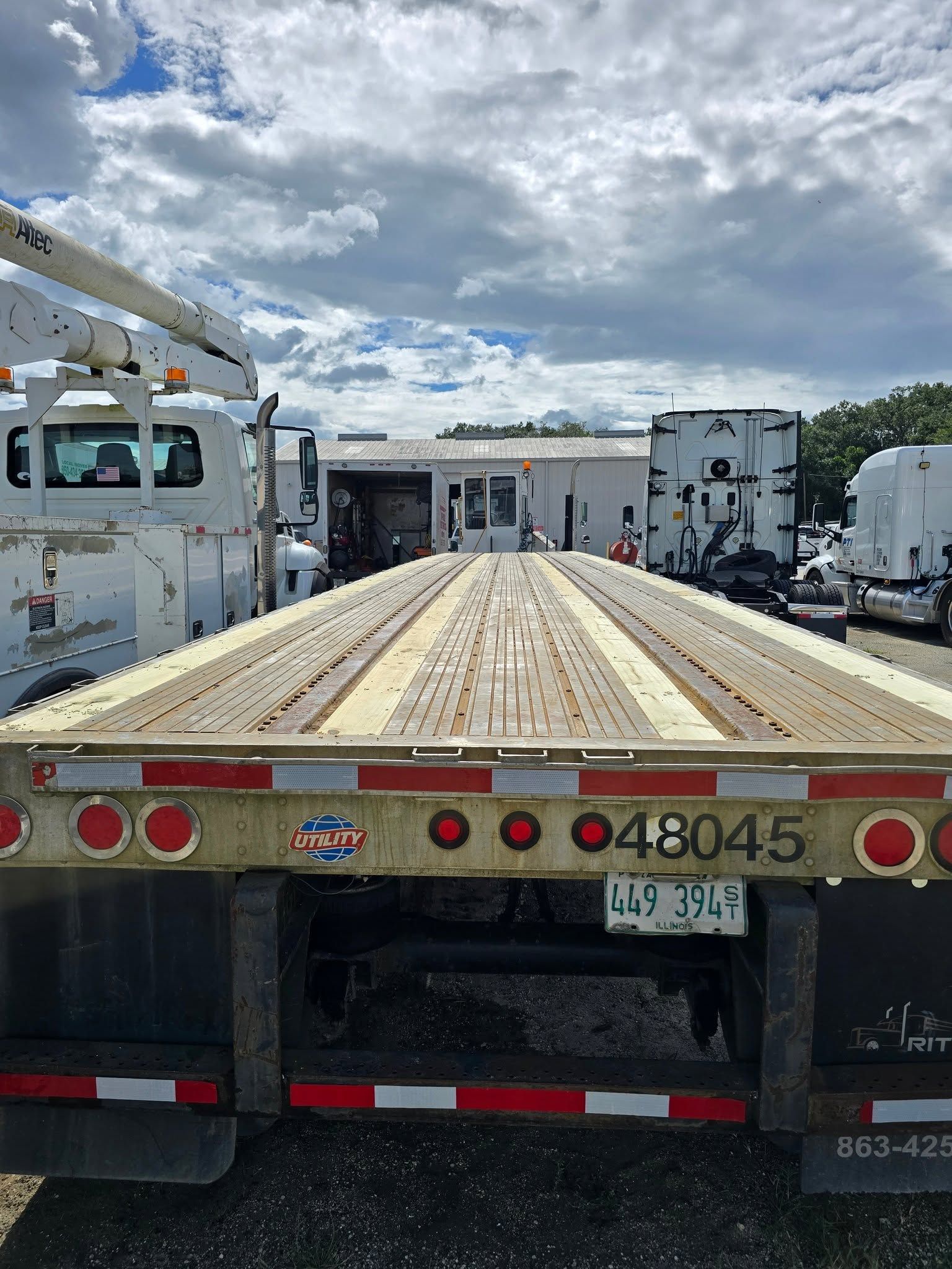 Flatbed tow truck carrying a long wooden platform under a cloudy sky on a roadside.