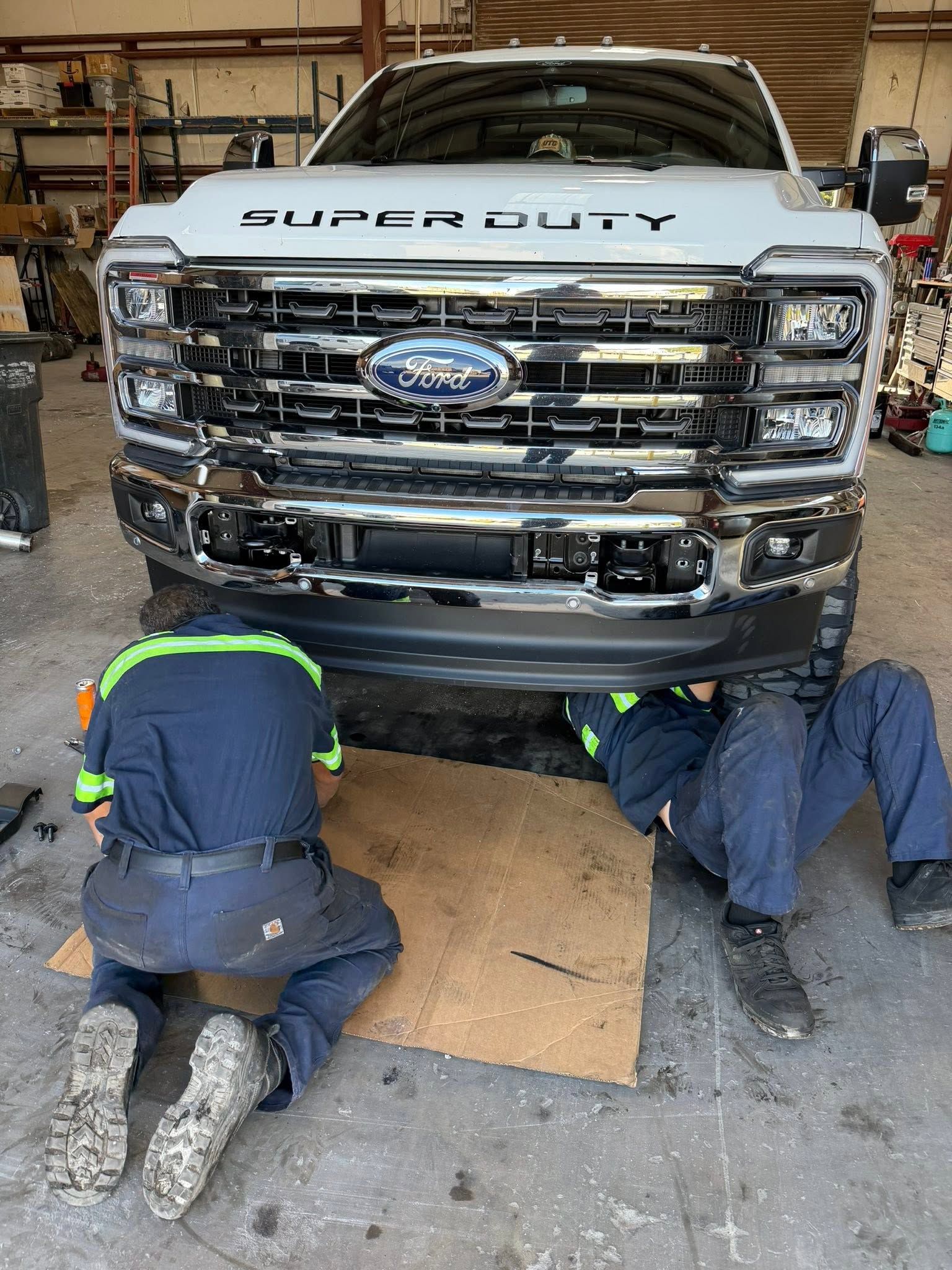Two mechanics working under the front of a white Ford Super Duty truck in a garage