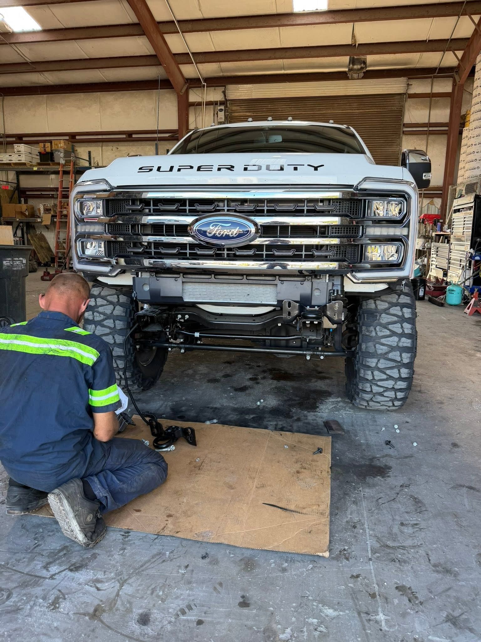 Mechanic working under a lifted Ford truck in a garage, front grille removed