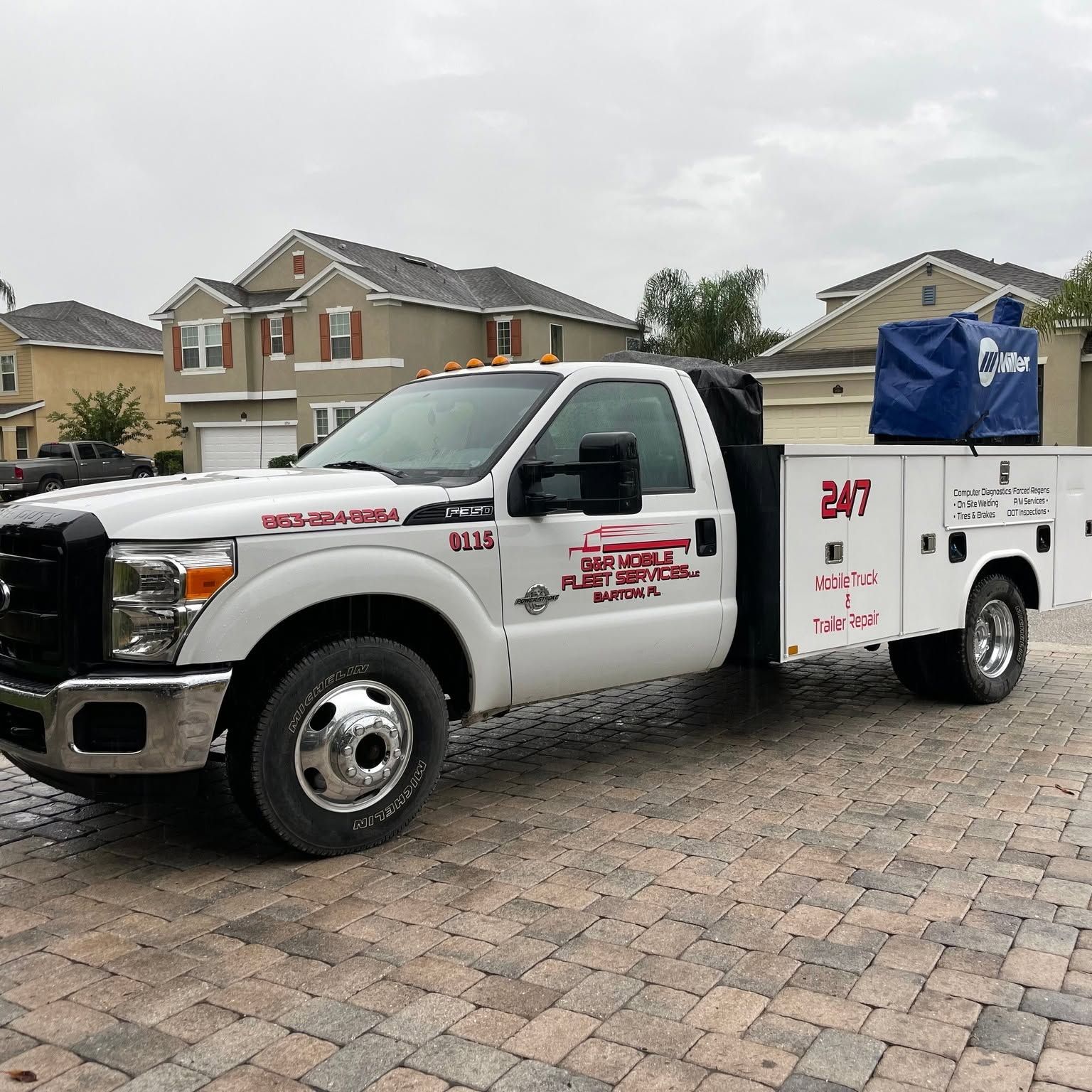 White tow truck with blue equipment parked on a brick driveway in a suburban neighborhood.