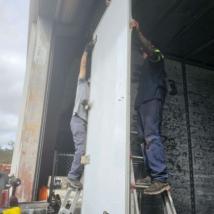 Two workers on ladders install a large white panel inside a truck trailer.