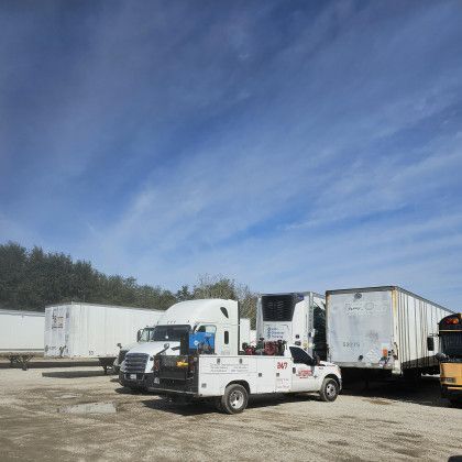 Trucks and trailers parked in a lot under a blue sky, including a white tow truck in front.