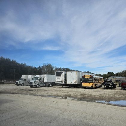 Parked trucks and trailers in a gravel lot under a cloudy blue sky