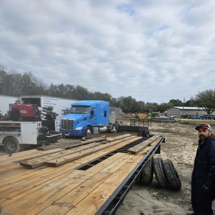 Flatbed trailer with lumber and a blue semi truck at a worksite, with a man standing nearby.