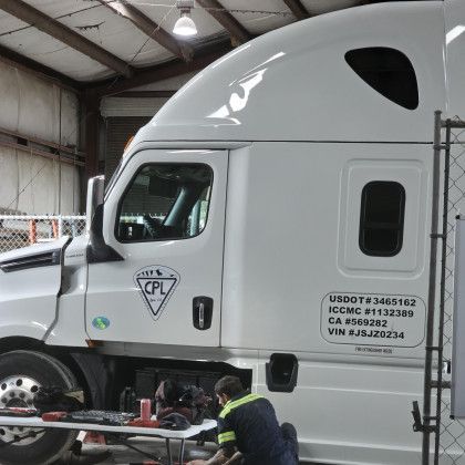 White semi-truck in a warehouse while a worker inspects the lower side of the cab.