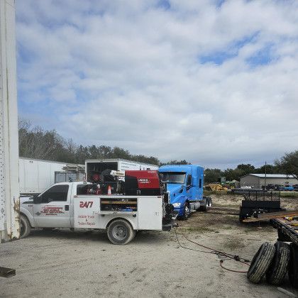 White tow truck with red number 847 and blue semi cab in a muddy truck yard under cloudy sky