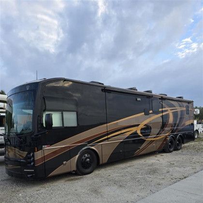 Large black and tan luxury motorcoach parked on a gravel lot under a cloudy sky