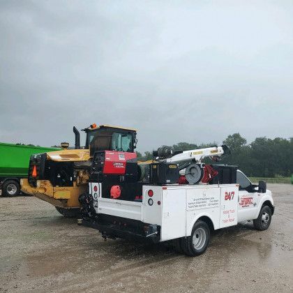 Tow truck hauling a large yellow construction machine in a wet gravel lot under cloudy skies