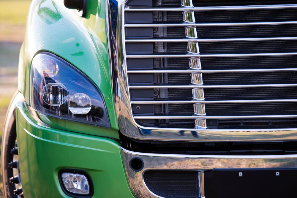 Close-up of a green truck front with chrome grille and headlight on a sunny street
