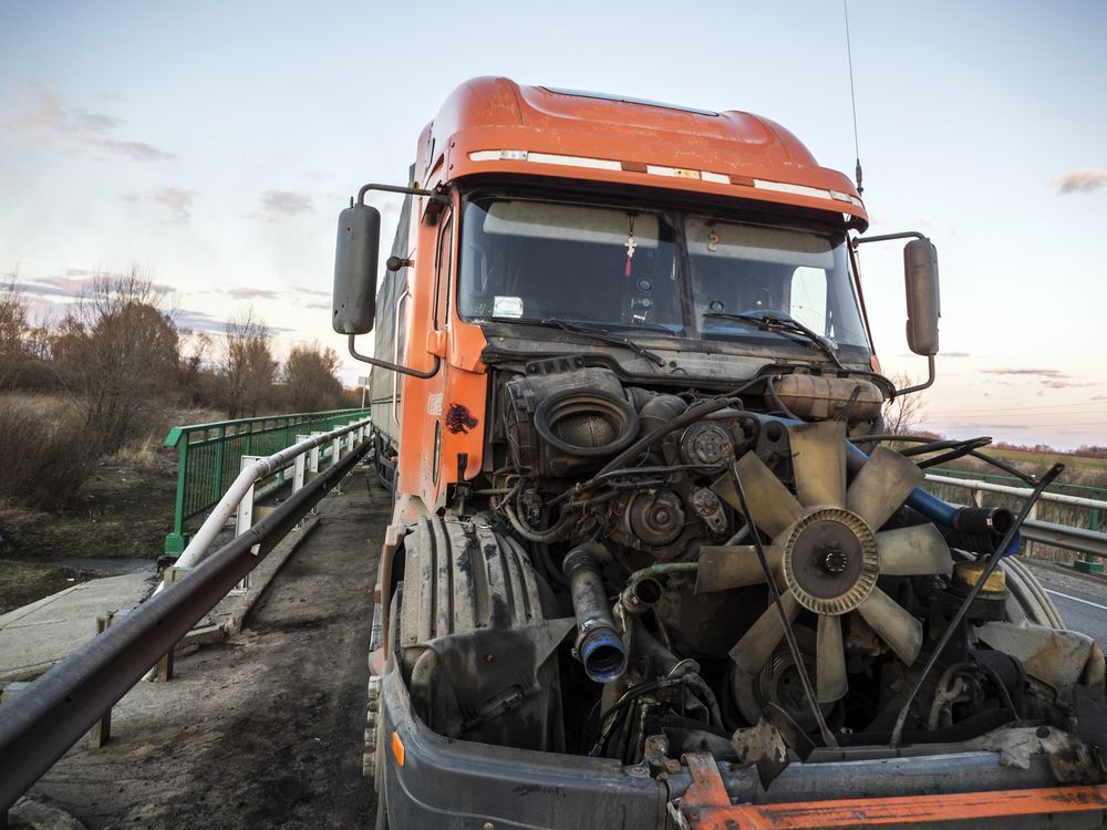 Orange truck with a smashed front end and exposed engine on a roadside bridge