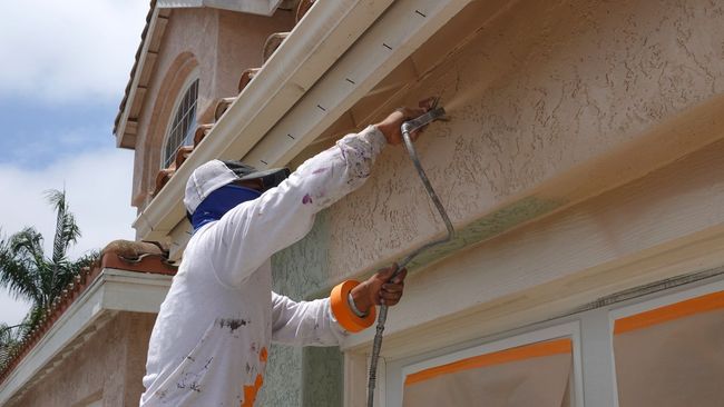 Worker painting the exterior trim of a house with a roller on a long extension pole Worker painting the exterior trim of a house with a roller on a long extension pole