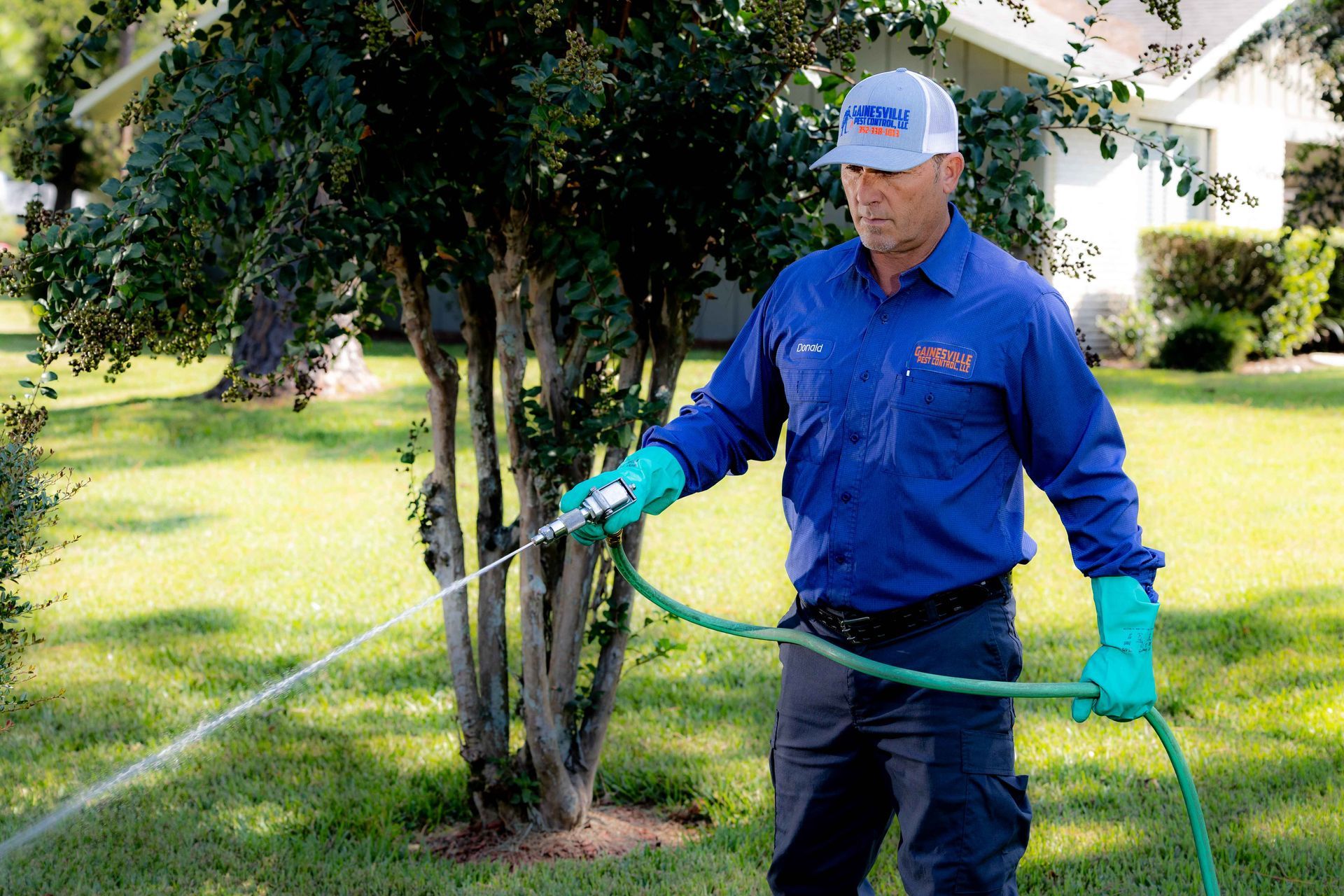 Pest control worker spraying lawn with hose. He wears blue uniform, gloves, and hat, standing outdoors.
