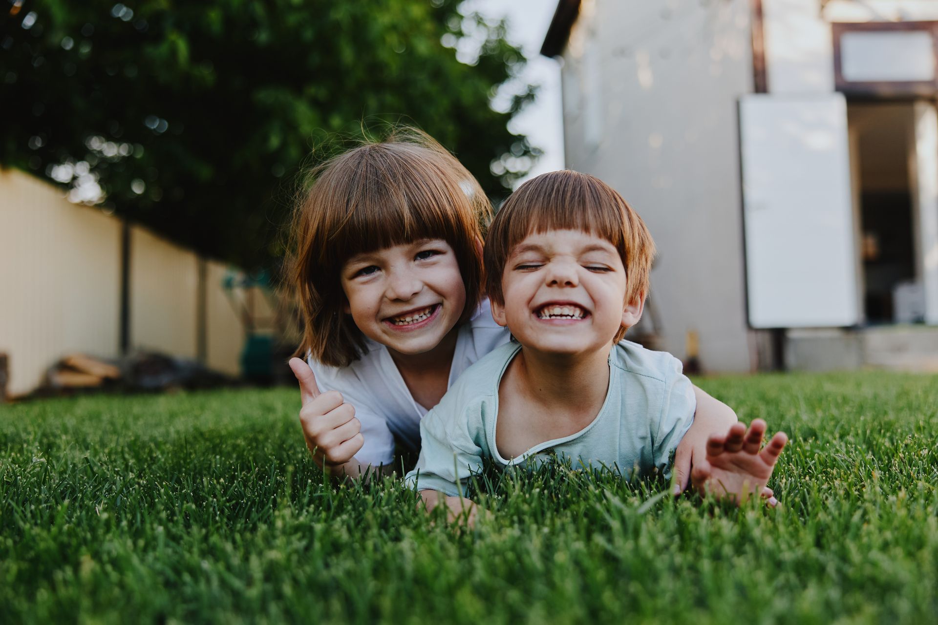 Two children smiling and lying in green grass outside a house
