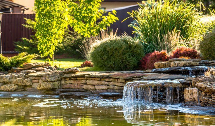 Waterfall cascading into a pond surrounded by tiered stonework and lush green landscaping.