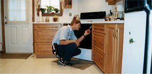 Man fixing oven in a kitchen with light wood cabinets and a white refrigerator.