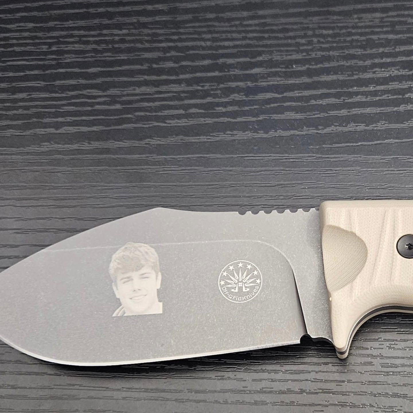 Gray knife with a serrated edge and a small engraved portrait on the blade, resting on a dark wood surface
