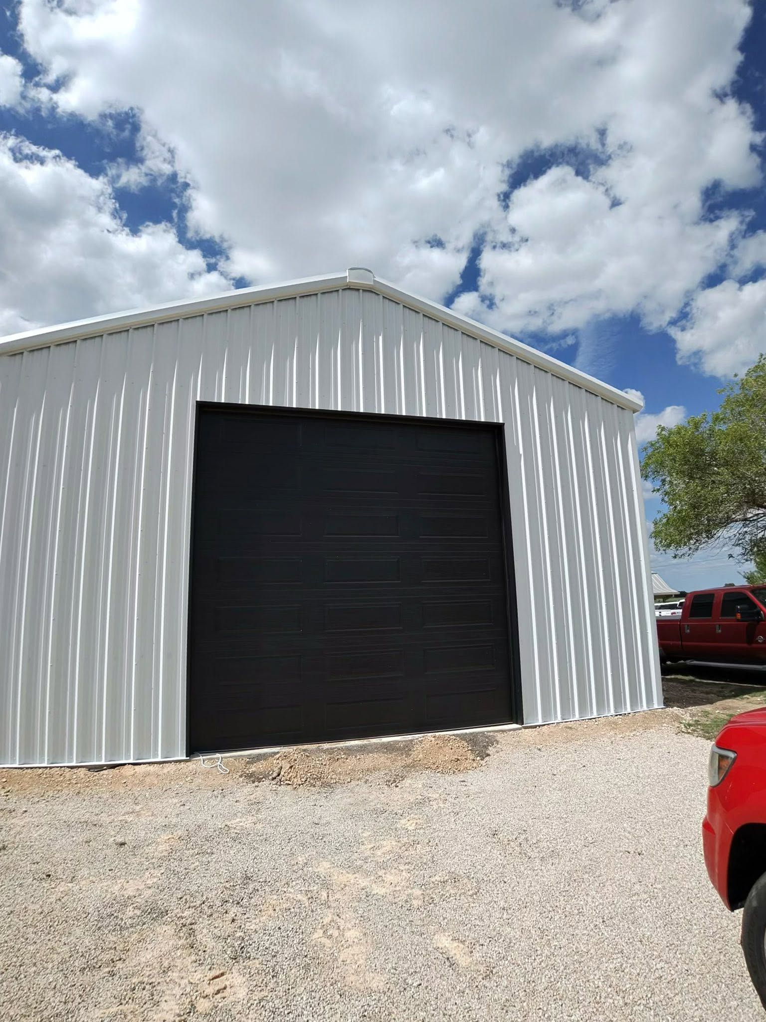 White metal barn with large black door on a gravel lot under a partly cloudy sky