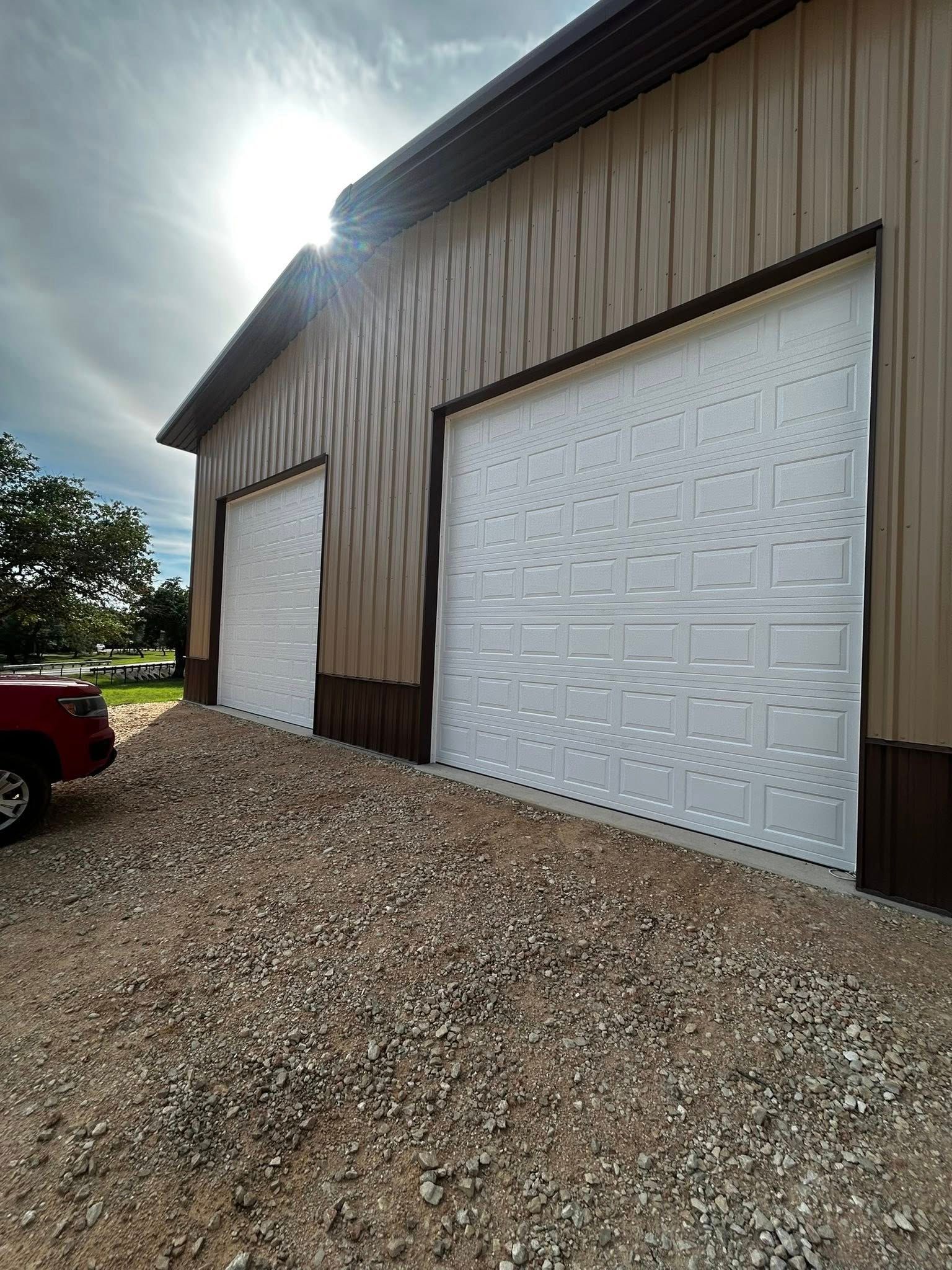 Large tan metal garage with white doors on a gravel driveway in bright sunlight
