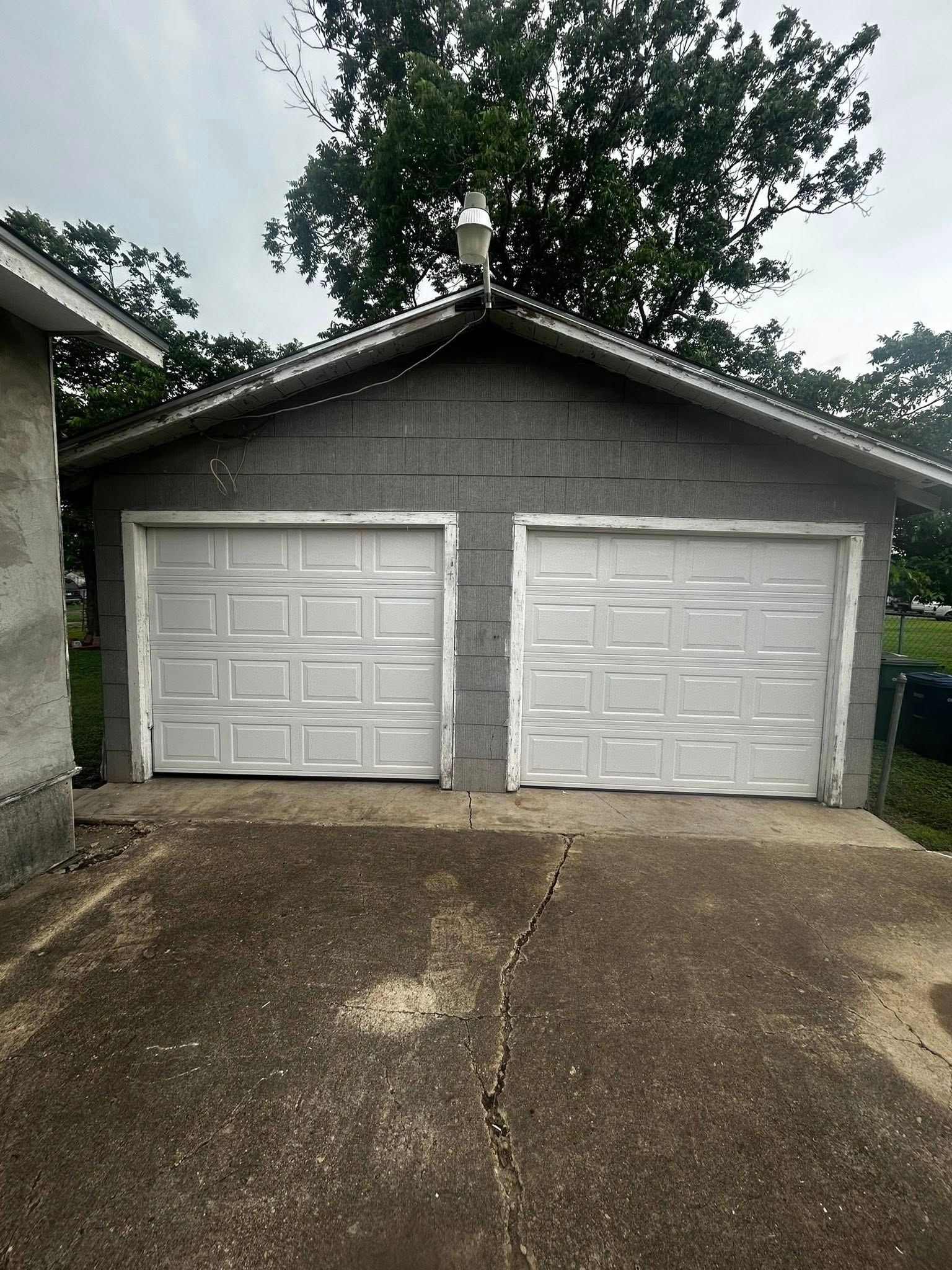 Two-door gray garage with white doors and a wet driveway under trees
