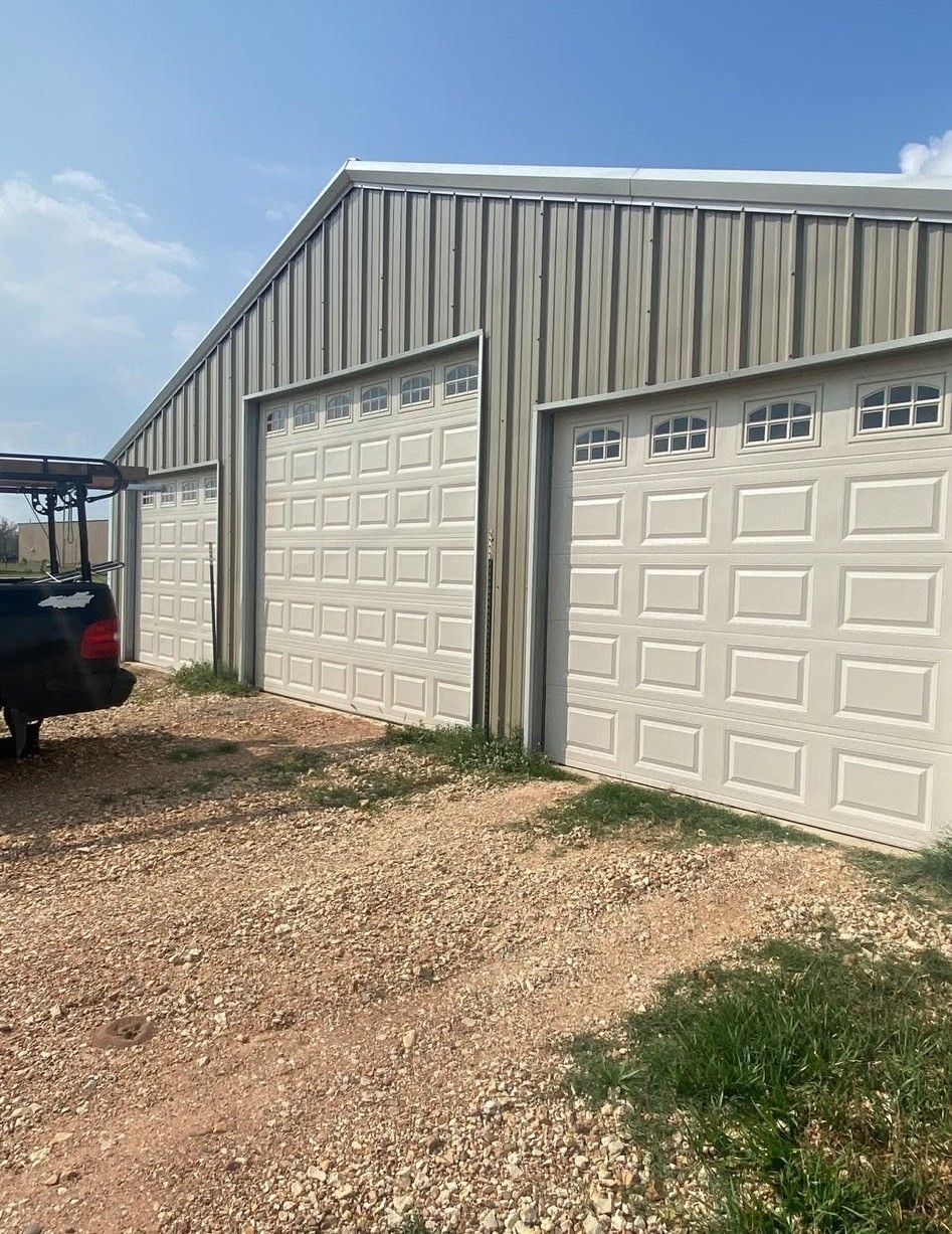 Three white garage doors on a metal building beside a gravel driveway under a blue sky
