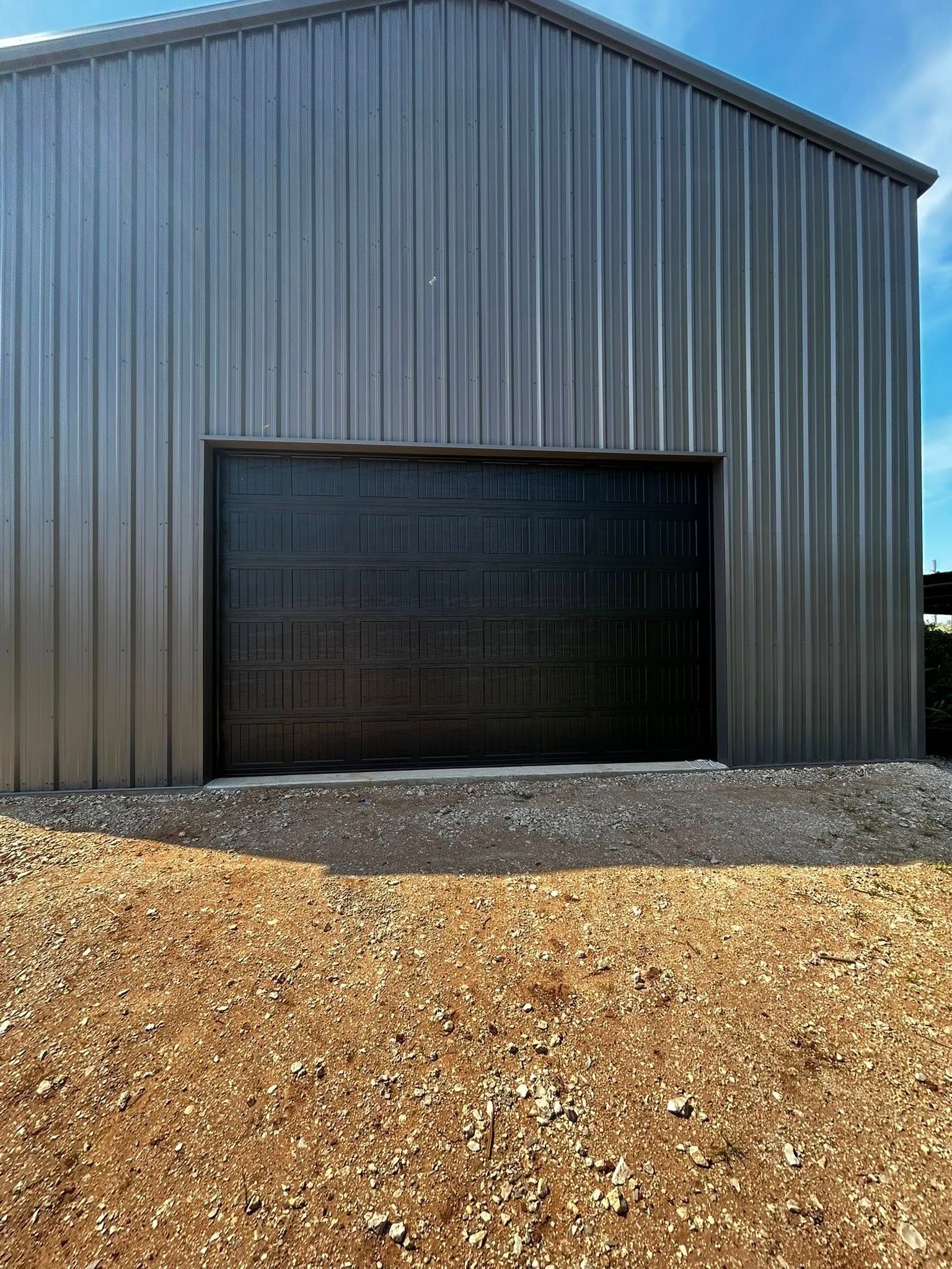 Gray metal garage building with a large black door and gravel foreground