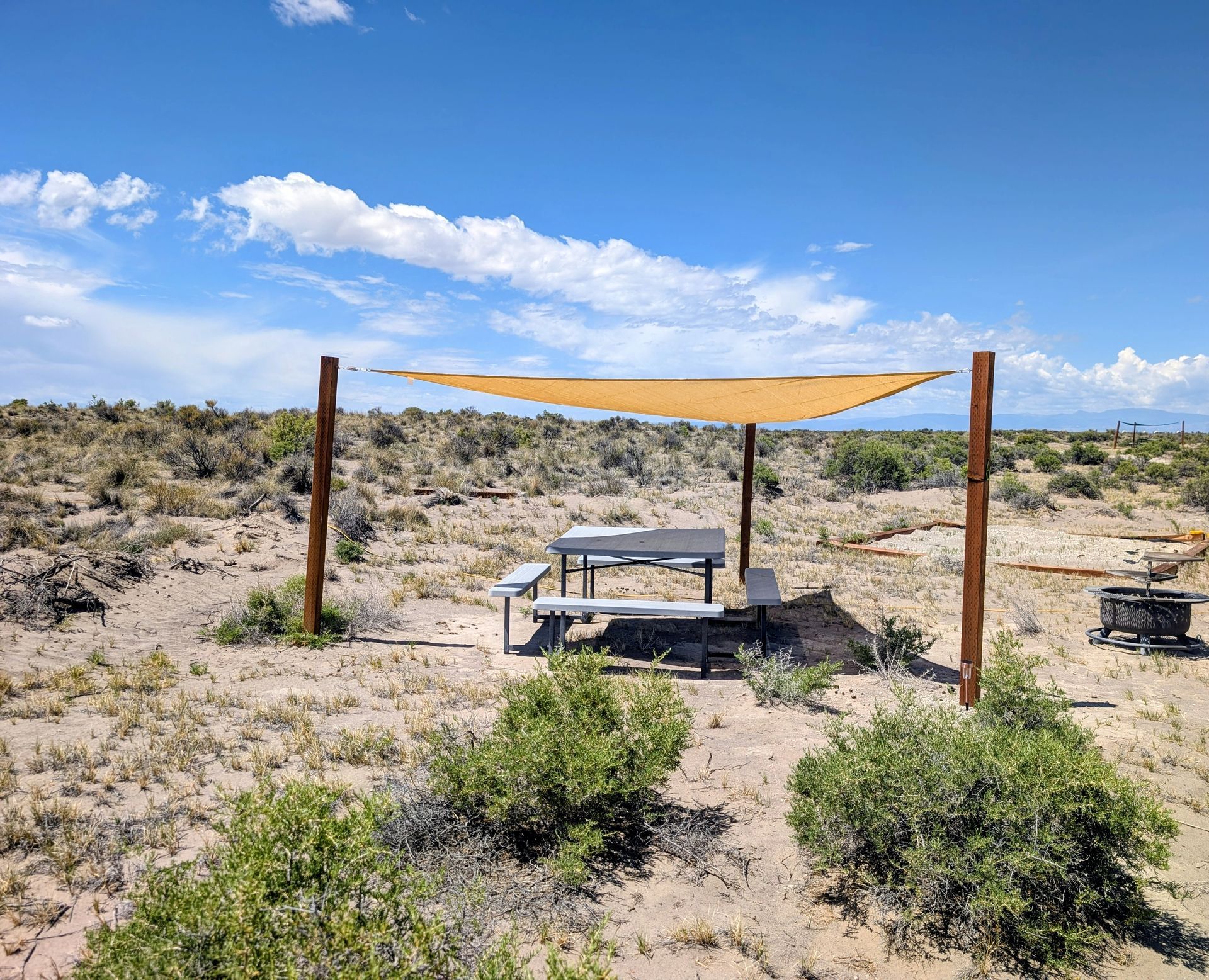 Desert picnic area with a shaded table under tan canopy, surrounded by scrub and blue sky.