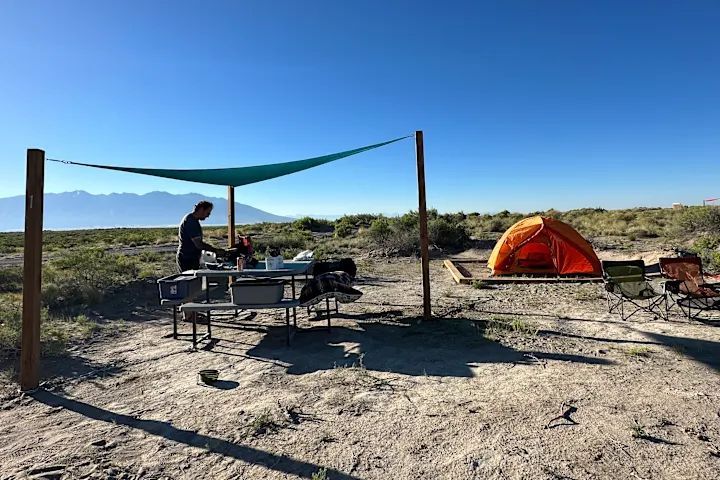 Desert campsite with a person at a picnic table, orange tent, and shade tarp under a clear blue sky