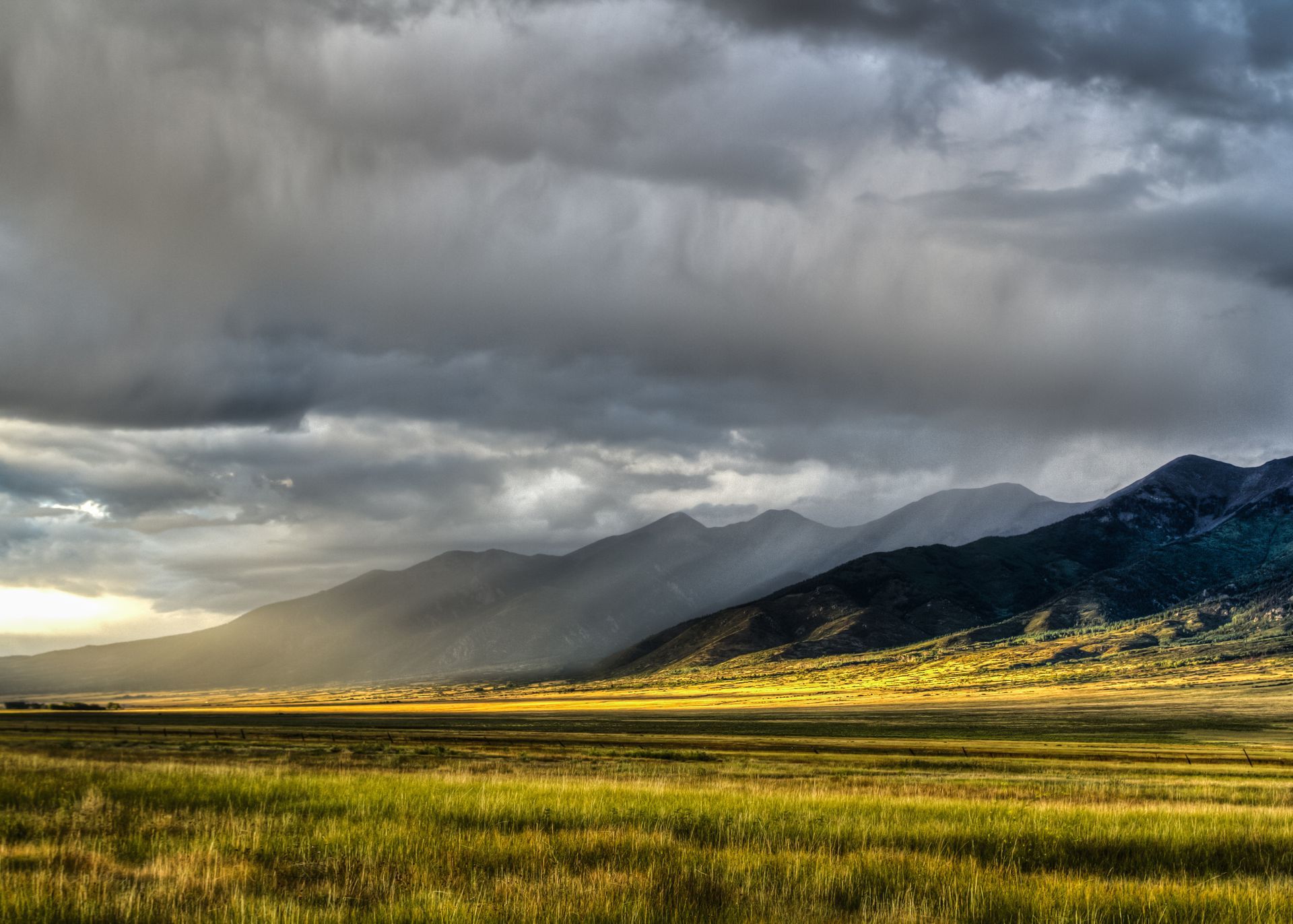 Sunlight on a golden field beneath dark storm clouds and a mountain range