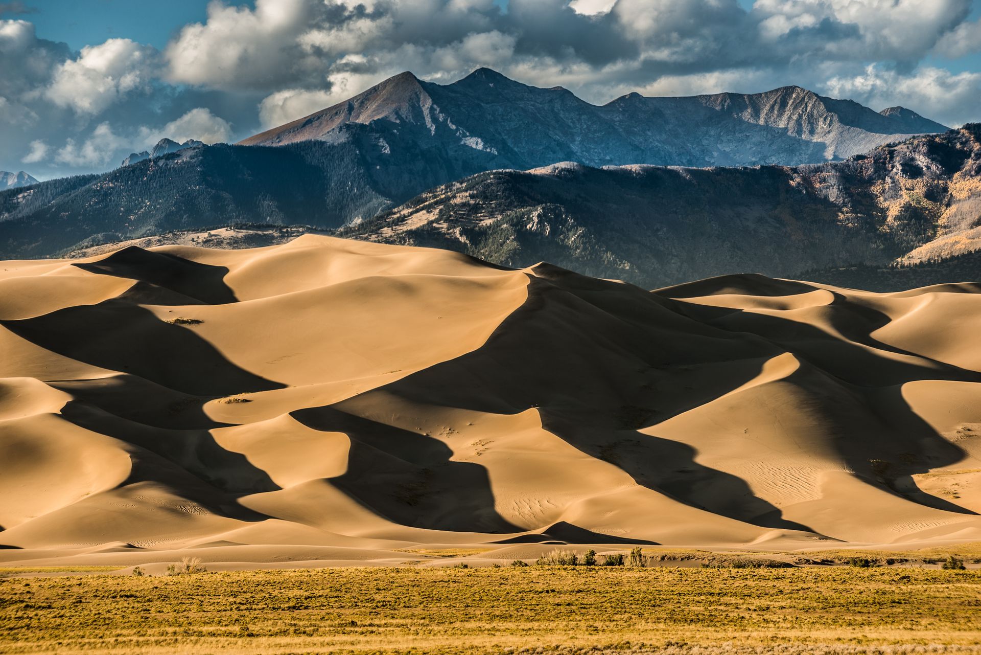 Sandy dunes with dramatic shadows before a rugged mountain range under a clear blue sky