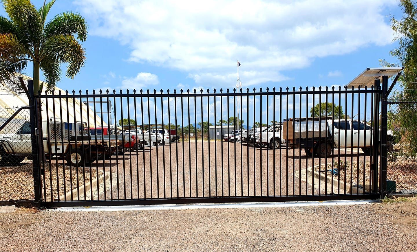 Black metal gate with vertical bars, open, revealing a gravel parking lot with parked vehicles under a blue sky — Gate-O-Matic in Garbutt, QLD