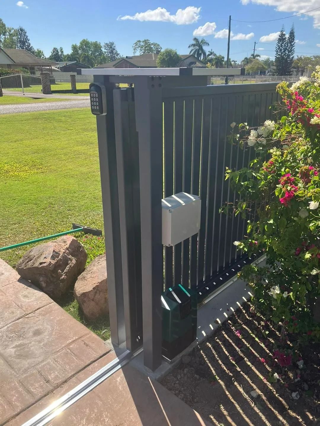 Black Metal Sliding Gate With Electronic Opener, Next to Green Grass and Flowering Bush — Gate-O-Matic in Garbutt, QLD