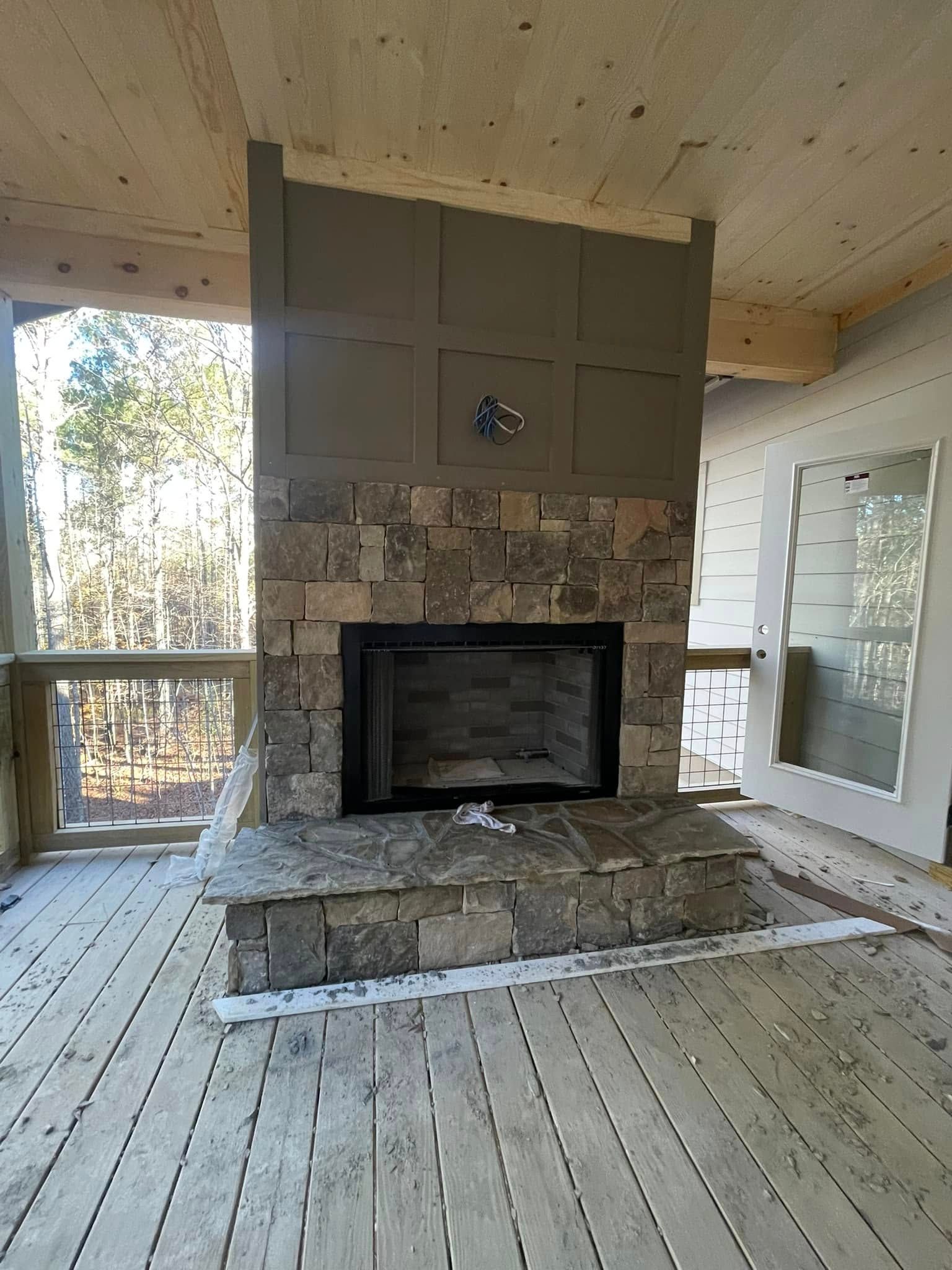A stone fireplace with a dark gray paneled upper section stands on a wood-planked outdoor deck overlooking a forest.