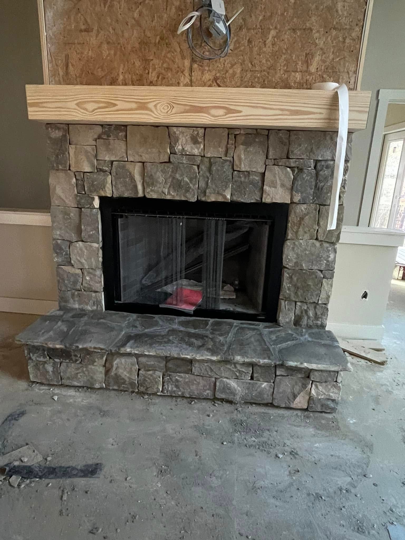 A stone fireplace under construction, featuring a natural wood mantel and a dark metal firebox in an unfinished room.