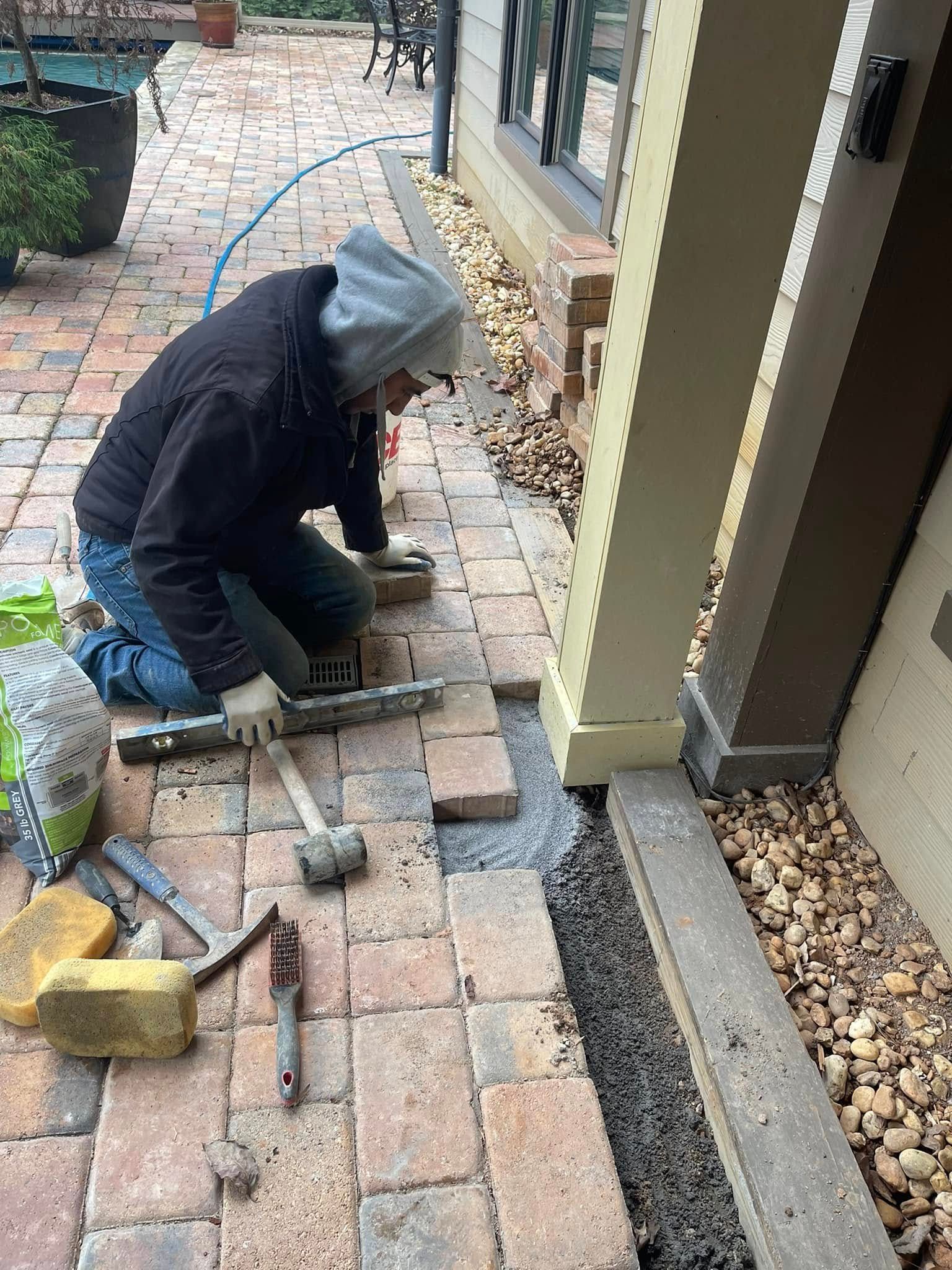 A person in a hooded jacket kneeling on a patio, using a rubber mallet and level to install brick pavers near a pillar.