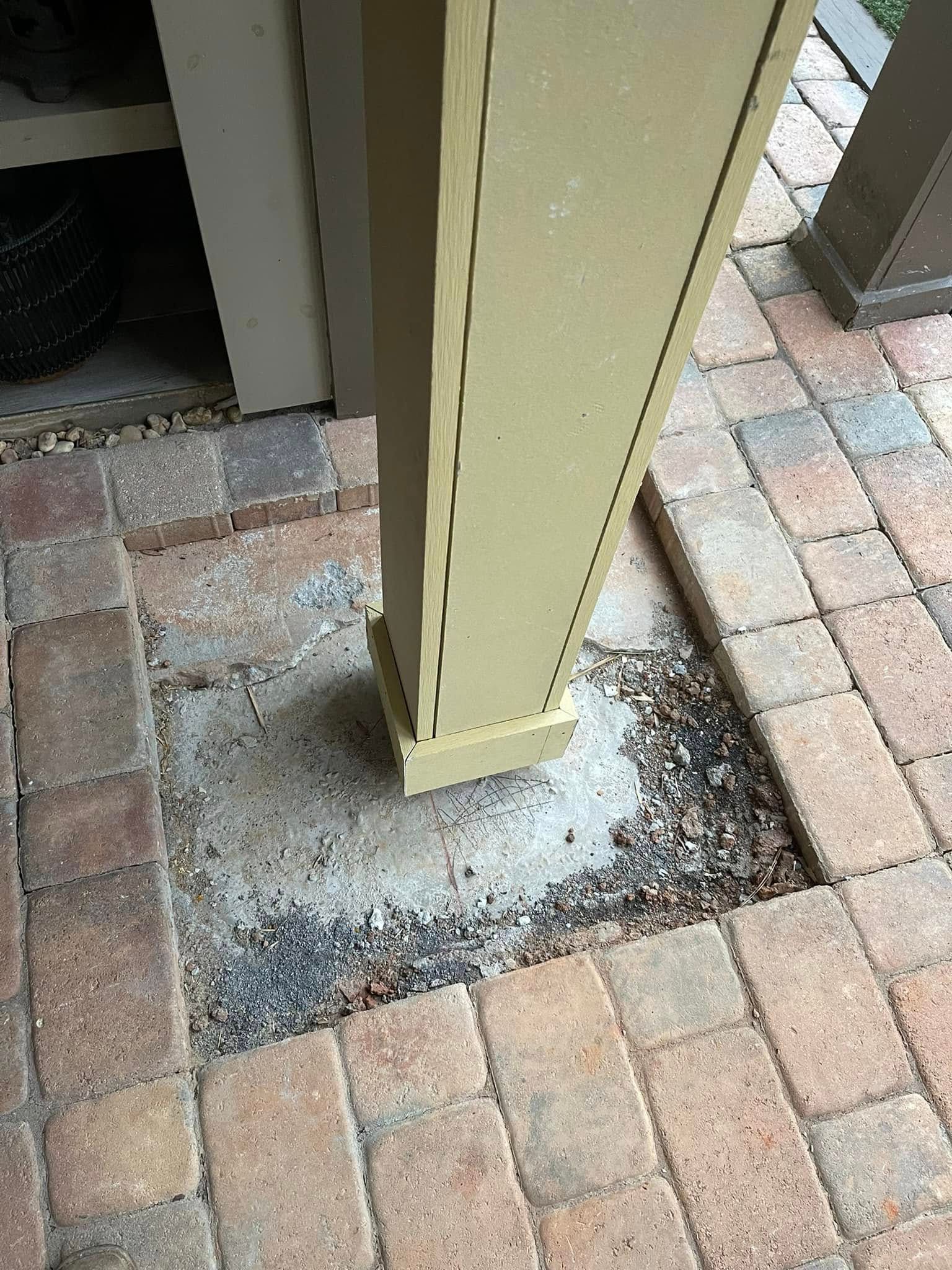 A beige wooden post base stands in the center of a square gap in a brick patio, exposing the underlying concrete footer.