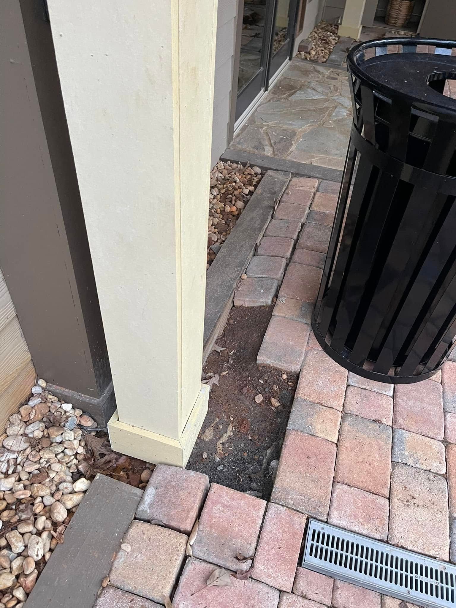 A light-colored structural post stands next to a gap in a brick paver walkway and a black outdoor trash receptacle.