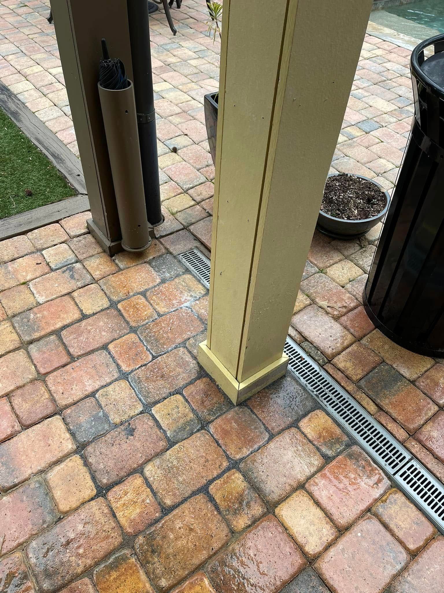 A cream-colored patio post stands on brick pavers next to a linear metal drainage grate and a black trash bin.
