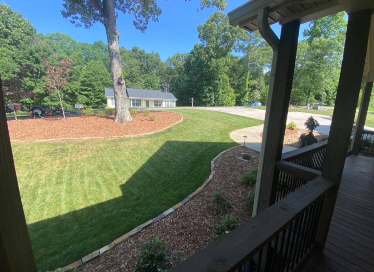 A view from a wooden porch looking out over a green lawn with landscaped mulch beds and a house in the distance.