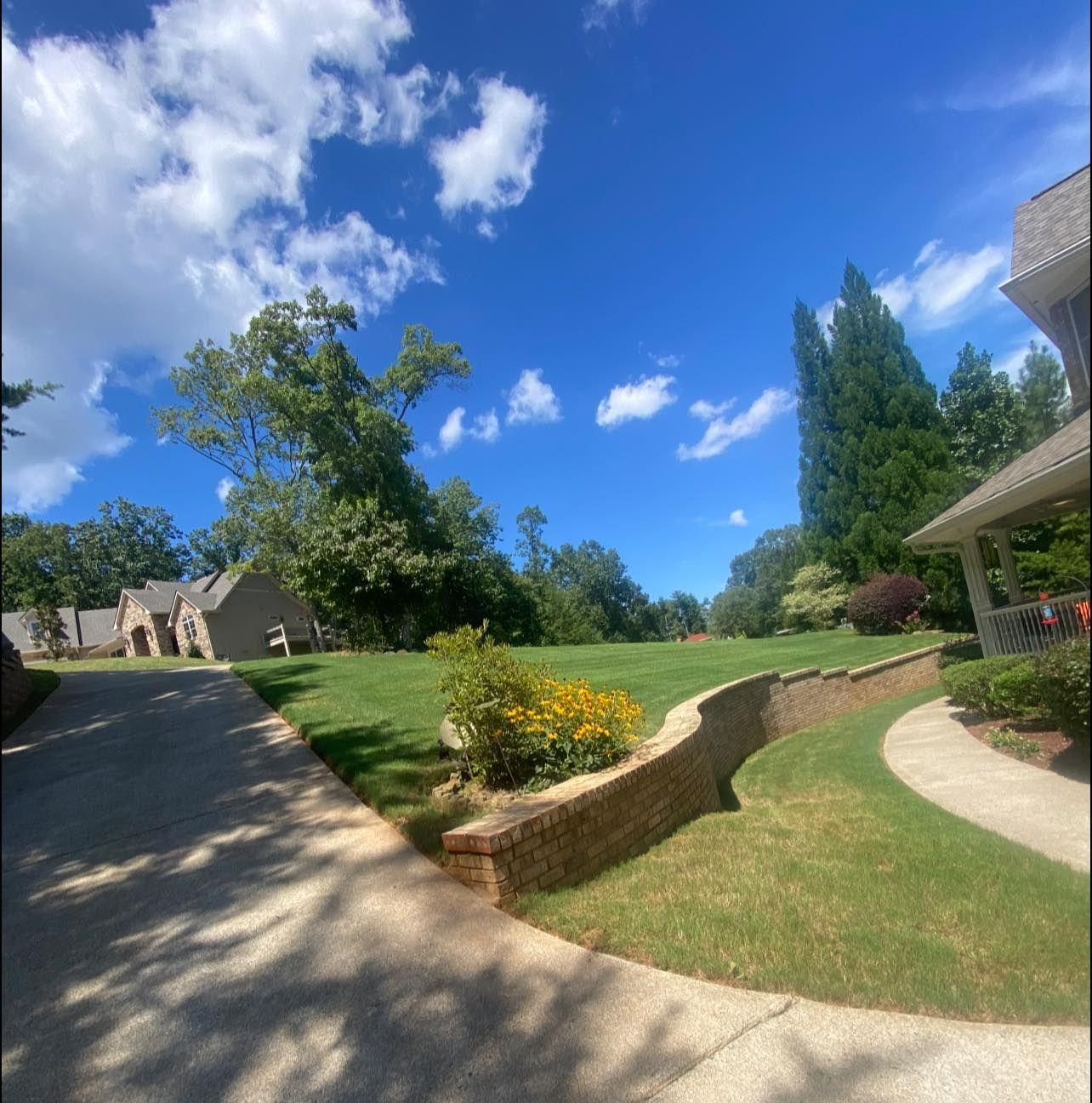 A scenic residential yard with a curved stone retaining wall, paved paths, manicured grass, and yellow flowers.
